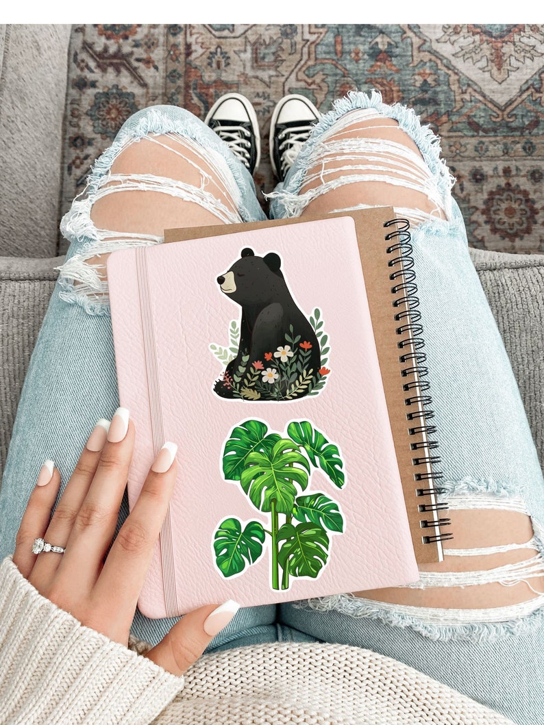 Puede incluir: Una libreta rosa con una pegatina de un oso negro y una planta de monstera verde. La libreta tiene una encuadernaci&oacute;n en espiral.