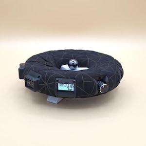 May include: A black, circular device with a geometric pattern, featuring a small screen displaying "BROADVIEW DESIGN". It has a silver ball on top and a control knob. The device is on a light beige surface.