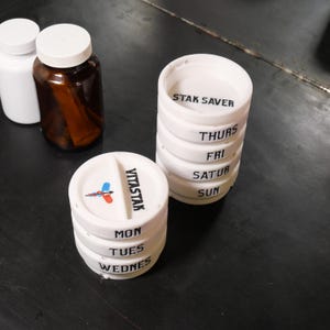 May include: White pill organizers with days of the week labeled, alongside two pill bottles. One organizer is labeled "STAK SAVER" and the other "VITASTAR". The organizers are cylindrical and have individual compartments for each day.