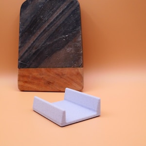 May include: A light purple, rectangular coaster with raised edges sits in front of a dark marble and wood cutting board. The cutting board has a dark gray and brown pattern.