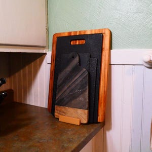 May include: A set of four cutting boards, including a black slate board, a brown wood board, and two smaller black boards, are stacked and leaning against a wall. The boards are held together by a wooden stand.