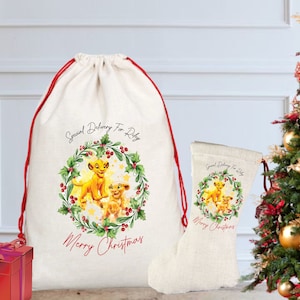 May include: A cream-colored Christmas gift sack and stocking featuring a watercolor illustration of lion cubs within a holly wreath. The sack has red drawstring and the text "Special Delivery For Riley" and "Merry Christmas". A red gift box and Christmas tree are in the background.