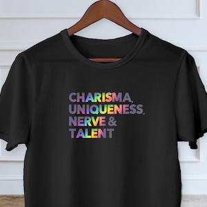 May include: Black t-shirt displaying the words "CHARISMA, UNIQUENESS, NERVE & TALENT" in a rainbow gradient. The tee is suspended on a wooden hanger, set against a white wall.