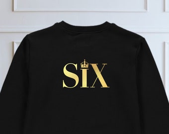 May include: Black sweatshirt with the word "SIX" in gold, featuring a crown above the "X". The design is centered on the chest. The sweatshirt is a solid black color.