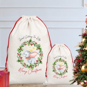 Puede incluir: Dos bolsas de regalo navideñas con cordón de color crema y ribete rojo. Cada bolsa presenta un elefante de dibujos animados dentro de una corona de acebo con el texto "Special Delivery For Riley" y "Merry Christmas". Una caja de regalo roja y un árbol de Navidad están en el fondo.