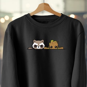 Puede incluir: Sudadera negra con un diseño de mapache de dibujos animados y Groot. El mapache tiene una expresión de enfado, y Groot es un personaje parecido a un árbol pequeño. La sudadera se muestra en una percha de madera.