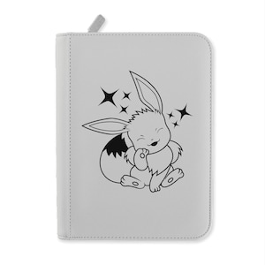 Puede incluir: Un estuche gris claro con cremallera con una ilustración en blanco y negro de una criatura parecida a un conejo de dibujos animados y estrellas brillantes. La criatura está sonriendo y tiene orejas grandes y una cola esponjosa. El estuche tiene un pequeño tirador gris.