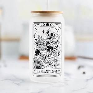 May include: White reusable tumbler with a black and white illustration of a skeleton holding a pot of plants. The text "THE PLANT LOVER" is printed below the illustration.