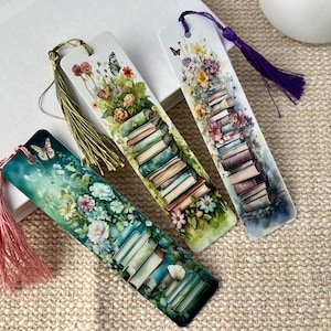 May include: Three colorful bookmarks with a stack of books, flowers, and butterflies. Each bookmark has a different color tassel.