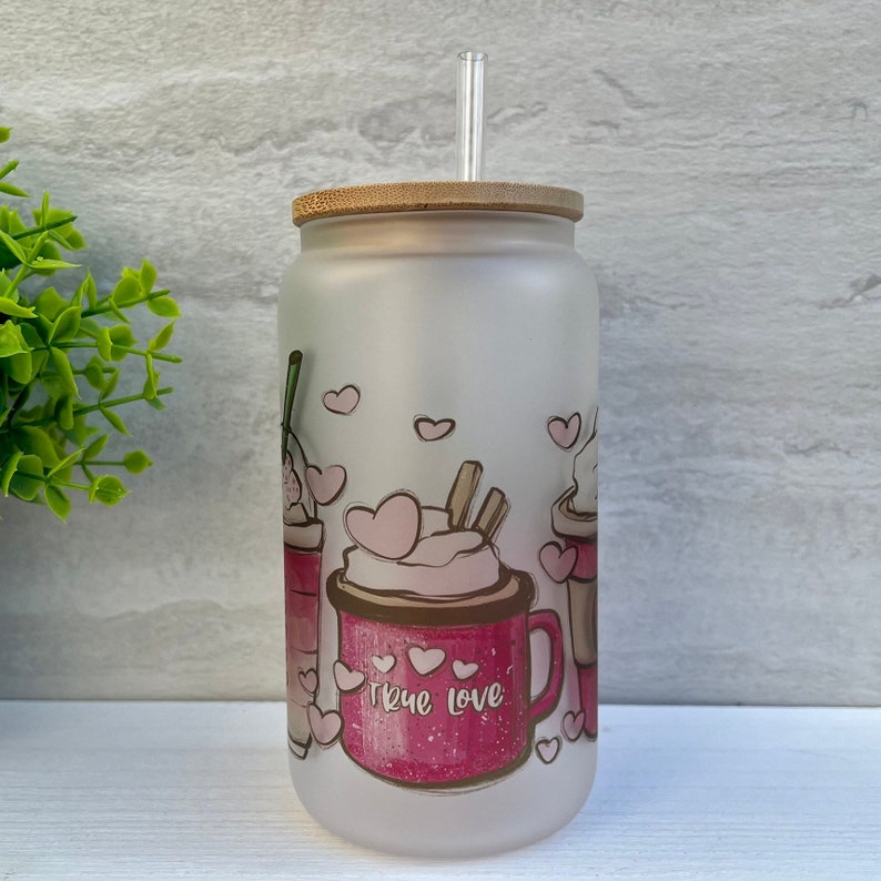 May include: A frosted glass tumbler with a bamboo lid and a clear straw. The tumbler has a pink and brown design of three coffee cups with whipped cream and hearts. The text "True Love" is printed on the front of the tumbler.