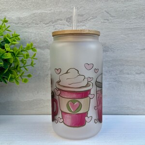 May include: A frosted glass tumbler with a bamboo lid and a clear drinking straw. The tumbler features a pink and white design of coffee cups with hearts and whipped cream.