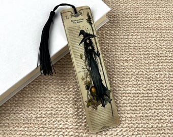 Può includere: Un segnalibro rettangolare con sfondo beige in stile vintage, caratterizzato da un'illustrazione a inchiostro nero di una strega con abito lungo e cappello a punta che tiene un bastone. Include testo stampato, decorazioni e una nappa nera.