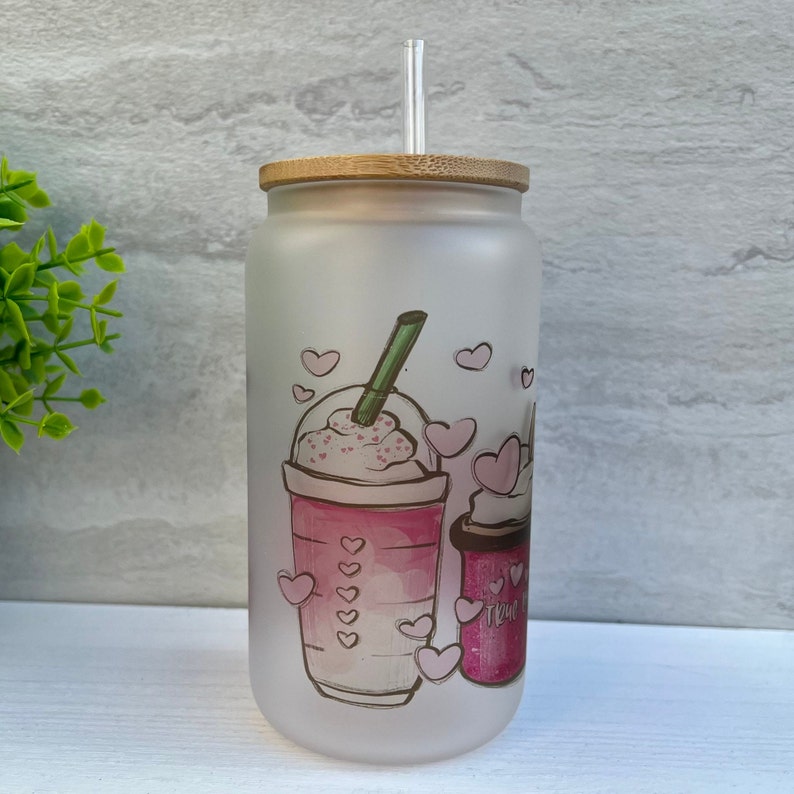May include: A frosted glass tumbler with a bamboo lid and a straw. The tumbler has a pink and white design of a coffee drink with whipped cream and hearts.