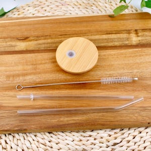 May include: A set of two clear glass drinking straws with a cleaning brush and a wooden lid. The lid is round and has a small hole in the center. The straws are on a wooden surface.