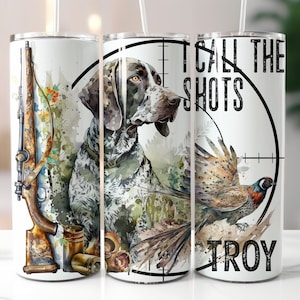 May include: A white tumbler with a hunting-themed design. The tumbler features a German Shorthaired Pointer dog, a shotgun, and a pheasant. The text "I call the shots" and "TROY" are printed on the tumbler.