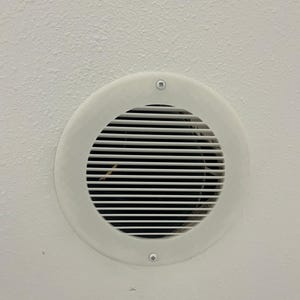Op de afbeelding: Een witte, ronde ventilatieafdekking met horizontale zwarte lamellen. De ventilatie is bevestigd aan een getextureerde, gebroken witte muur met twee kleine schroeven. De diameter is ongeveer 20 cm.