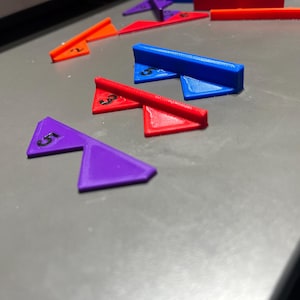 May include: A collection of brightly coloured, 3D-printed geometric shapes. The shapes include orange, red, blue, and purple triangles and rectangular bars. Each triangle has a number printed on it in black.