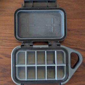 May include: A black and gray plastic storage case with a hinged lid and a handle. The case has a removable tray with 12 compartments.