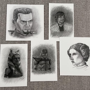 May include: Five pencil sketches of Star Wars characters on white paper. The sketches include portraits of Captain Rex, Luke Skywalker, Ahsoka Tano, a Clone Trooper, and Princess Leia. Each drawing is detailed with shading and highlights.