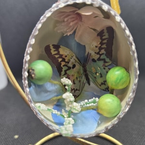May include: A delicate butterfly with green and brown wings is encased in a clear egg-shaped ornament. The ornament is decorated with a white and blue floral design and green beads.