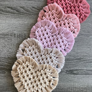 Puede incluir: Cinco posavasos de ganchillo en forma de corazón en tonos rosa y beige con detalles de flecos. Los posavasos están dispuestos en degradado de rosa oscuro a beige.