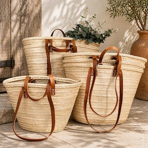 Pu&ograve; includere: Quattro borse tote in paglia intrecciata con manici in pelle marrone. Le borse variano di dimensioni, con la pi&ugrave; grande sul retro e la pi&ugrave; piccola sul davanti. Le borse sono vuote e poggiano su una superficie di pietra, con una porta di legno sullo sfondo.
