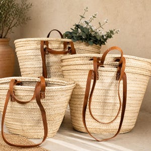 Pu&ograve; includere: Una collezione di borse tote in paglia intrecciata di varie dimensioni. Ogni borsa presenta manici e dettagli in pelle marrone. Le borse sono disposte insieme, alcune contengono vegetazione, suggerendo un'estetica naturale e rustica.