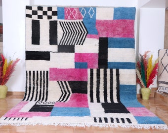 May include: Abstract geometric rug with a white background and black, pink, and blue geometric shapes. The rug has a fringe border.