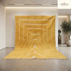 May include: A large, rectangular, mustard yellow rug featuring a series of concentric, nested square patterns in a lighter cream colour. The rug has a thick, plush texture and is finished with a short, knotted fringe along the bottom edge.