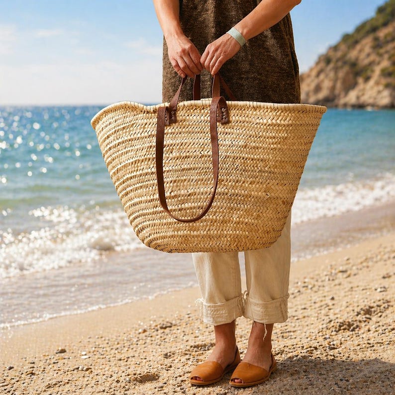 Pu&ograve; includere: Una grande borsa tote in paglia intrecciata con manici in pelle marrone. La borsa &egrave; tenuta da una persona su una spiaggia sabbiosa con l'oceano sullo sfondo. La borsa &egrave; di colore beige naturale e i manici in pelle sono marroni scuro.