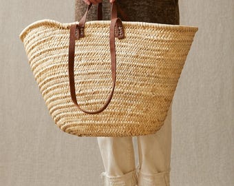 May include: A large, woven straw tote bag with brown leather handles. The bag is held by a person. The bag's open top and spacious interior are visible. The bag is made of natural-colored straw and has a classic, timeless design.