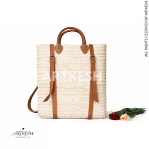 May include: A rectangular woven straw tote bag with brown leather handles and straps. The bag has a light beige colour and features the text "ARTKESH". A small bouquet of flowers rests near the bag.