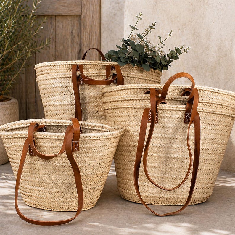 Pu&ograve; includere: Una collezione di cesti in paglia intrecciata di varie dimensioni, ciascuno con manici in pelle marrone. I cesti hanno un colore naturale, marrone chiaro e sono disposti insieme. Alcuni cesti contengono vegetazione.