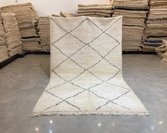 May include: Cream-colored, rectangular rug with a diamond pattern in black lines. The rug has a thick, textured pile and a fringed edge. The rug is displayed on a concrete floor.
