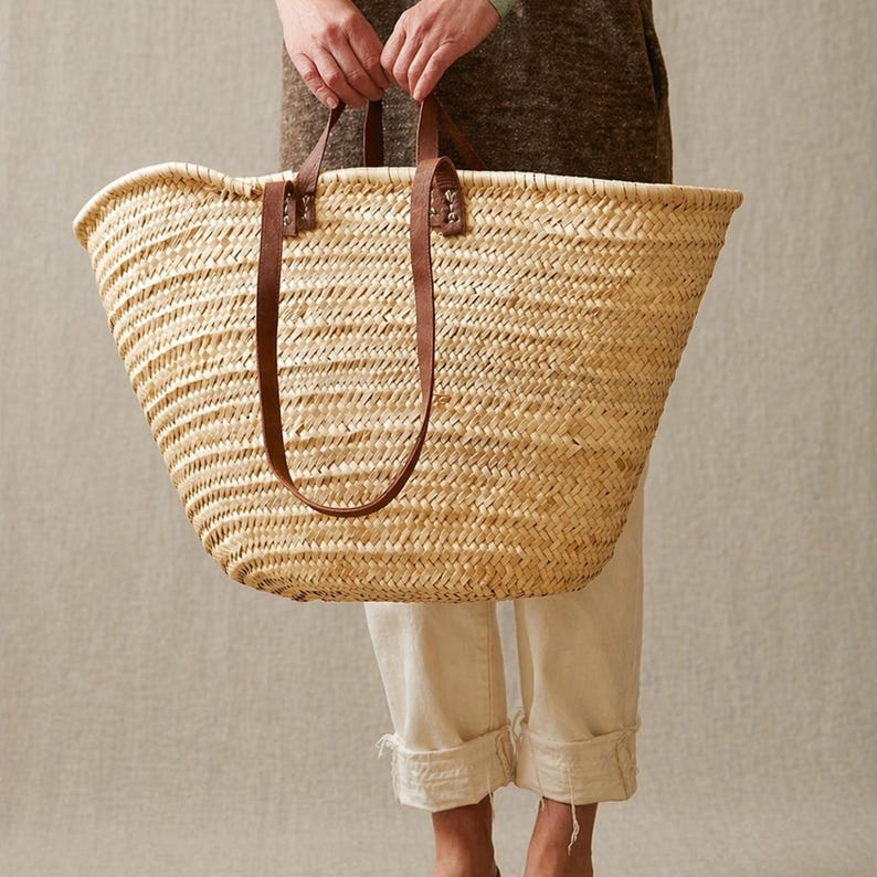Pu&ograve; includere: Una grande borsa tote in paglia intrecciata con manici in pelle marrone. La borsa &egrave; tenuta da una persona. Sono visibili la parte superiore aperta e l'interno spazioso della borsa. La borsa &egrave; realizzata in paglia color naturale e ha un design classico e senza tempo.