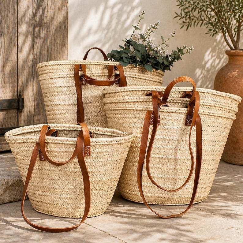 Pu&ograve; includere: Quattro borse tote in paglia intrecciata con manici in pelle marrone. Le borse variano per dimensioni e forma, con un colore beige naturale. I manici sono attaccati con cuciture visibili. Le borse sono disposte su una superficie di pietra.