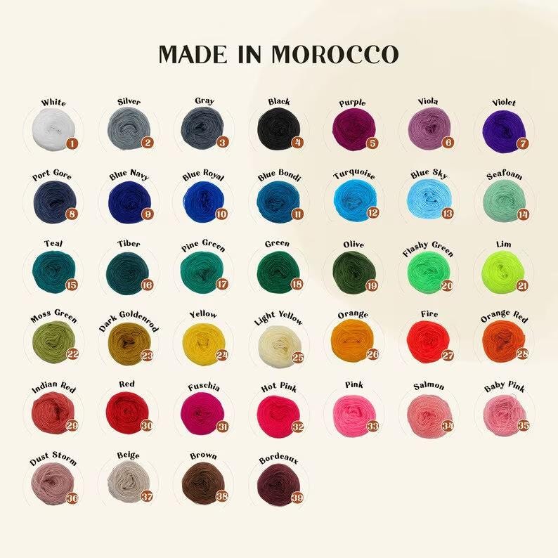 Pu&ograve; includere: Una tabella colori di gomitoli di filato in vari colori, tra cui bianco, nero, viola e verde. Il testo "MADE IN MOROCCO" &egrave; in alto. Ogni colore &egrave; etichettato con un nome e un numero.
