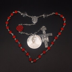 May include: A red coral rosary with a silver crucifix, a silver medal with the image of St. Padre Pio, and a red rose charm.
