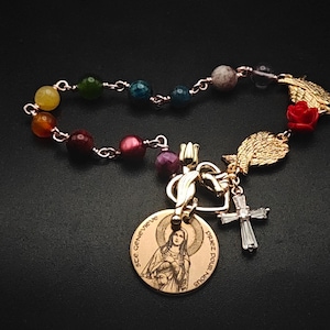 May include: A bracelet featuring a series of multicoloured beads connected by gold-toned metal links. It includes a round gold-toned medal depicting a figure with the text "Ste. Genevieve Priez Pour Nous," a gold-toned cross charm, a red rose, and gold wings.