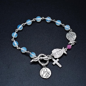 Op de afbeelding: Een zilveren armband met lichtblauwe kralen en religieuze bedels. De bedels omvatten een kruis, engelen vleugels en een medaillon met een religieuze figuur. De armband staat tegen een donkere achtergrond.