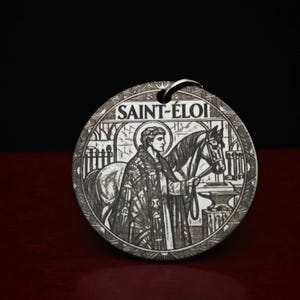 Op de afbeelding: Een ronde metalen hanger met een gegraveerde afbeelding van Sint-Elooi die naast een paard staat. Het ontwerp bevat een aambeeld, gereedschap en de tekst SAINT-ÉLOI bovenaan. De hanger heeft een zilverkleurige afwerking met donkere etsdetails.