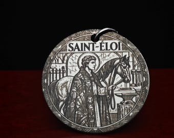 Può includere: Un ciondolo circolare in metallo con un'illustrazione incisa di Sant'Eligio in piedi accanto a un cavallo. Il design include un'incudine, attrezzi e il testo SAINT-ÉLOI in alto. Il ciondolo ha una finitura color argento con dettagli incisi.