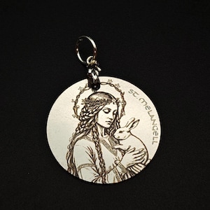 May include: A silver-coloured circular pendant with an engraved image of a woman holding a rabbit, with the text "St. Melangell" around the edge. A key ring is attached at the top.