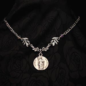 Op de afbeelding: Zilverkleurige ketting met een hanger met een afbeelding van een heilige en de tekst "SAINTE WALBURGE". De ketting heeft blad- en kristalaccenten, tegen een donkere bloemenachtergrond.