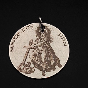May include: A circular, silver-toned pendant featuring an engraved image of a saint with a cross and staff. The text "SAINTE-FOY p.p.N" is around the edge. A small metal ring is attached for suspension.