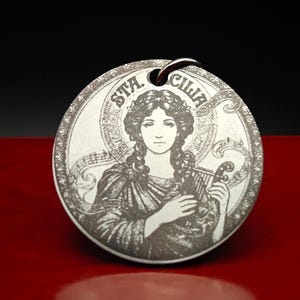 Peut inclure: Un pendentif circulaire en métal argenté avec une illustration gravée au laser représentant une figure portant une couronne de fleurs et tenant une petite harpe. Le texte STA. CILIA est inscrit en haut, avec des notes de musique en arrière-plan.