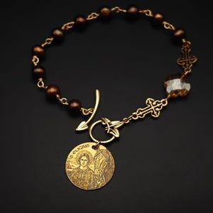 May include: A bracelet featuring dark brown beads, gold-toned metal links, and two ornate cross charms. A toggle clasp secures the piece, which includes a round gold-toned pendant engraved with an image of a person and a horse, labelled S. ELIGIUS.