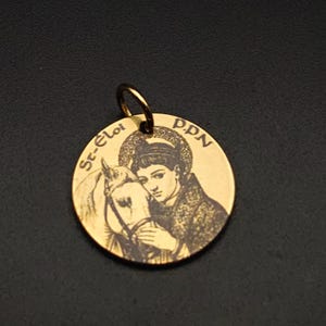 Peut inclure: Un pendentif doré de forme circulaire. Il présente une image gravée d'une personne avec un cheval, entourée des inscriptions "St-Eloi" et "D.P.N.". Le pendentif est doté d'une petite boucle pour une chaîne.