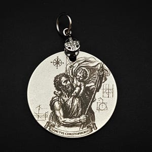 May include: A circular silver-toned pendant featuring an engraved illustration of Saint Christopher carrying the Christ child. The pendant includes a compass rose, geometric symbols, and the text SANCTUS CHRISTOPHORUS, topped with a small bail.