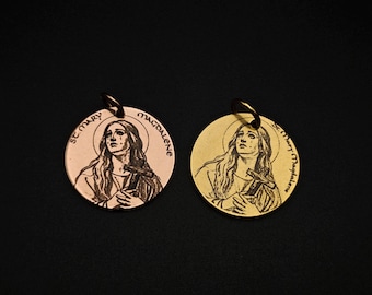 May include: Two round pendants featuring an engraved image of St. Mary Magdalene holding a cross. One pendant is copper-colored, the other is gold-colored. Both pendants have a small loop at the top for hanging.